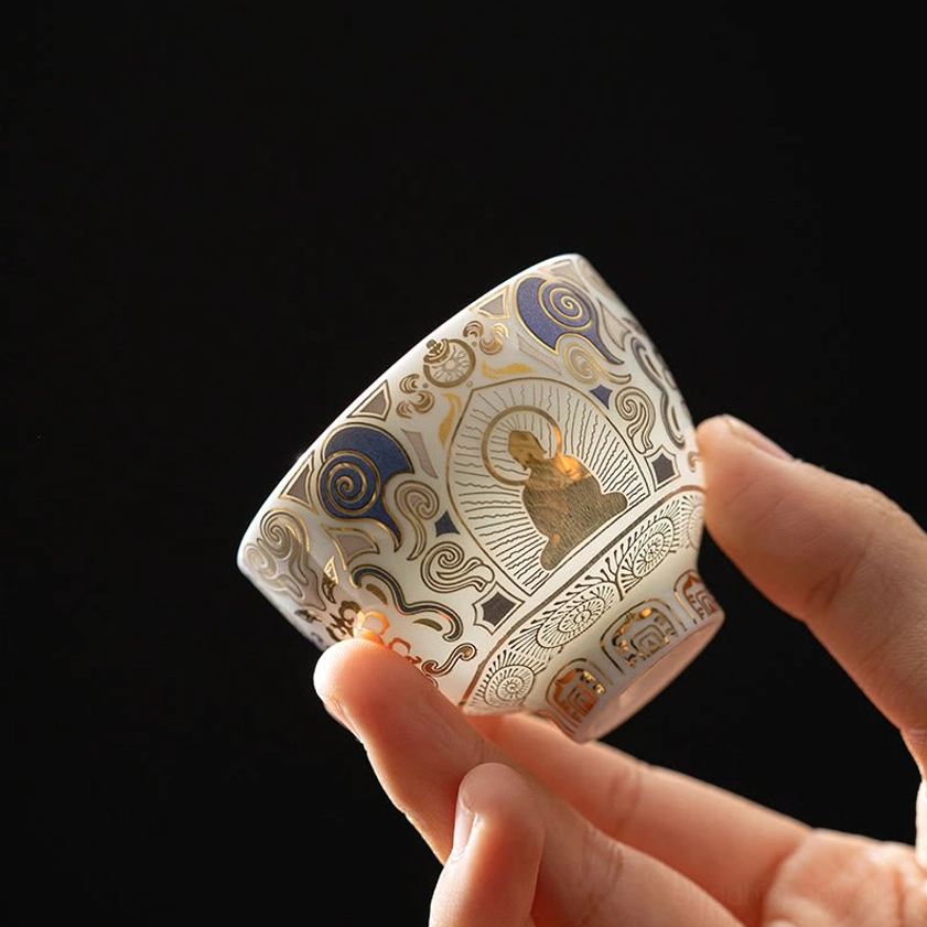 Handcrafted white ceramic kung fu tea cup with gilded Buddha and auspicious clouds design, held in hands against dark studio background, showcasing side view of Buddhist teacup