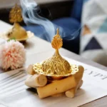 Gold Buddhist incense burner with smoking incense cone and wooden coaster on home table with flowers - top three-quarter view