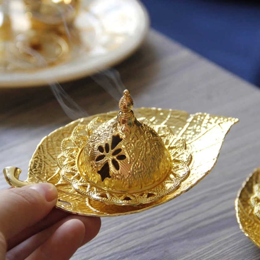 Hand holding gold incense burner with smoking incense cone, displaying detailed hollow carvings and golden metal
