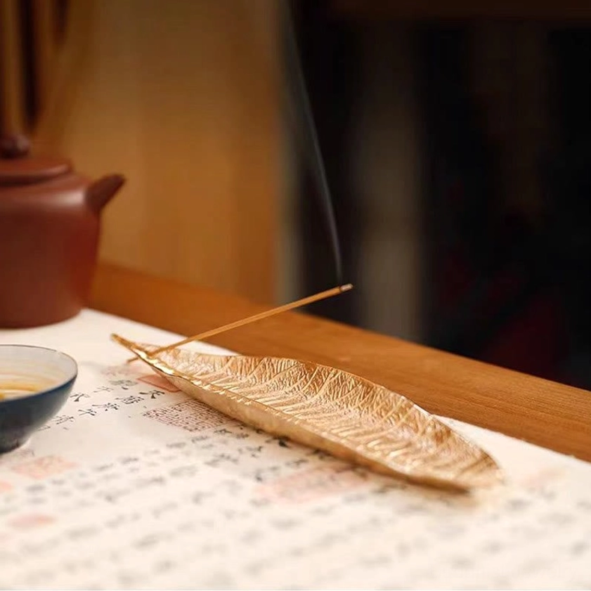 Golden leaf incense holder with lit incense and rising smoke beside teapot on calligraphy paper table setting