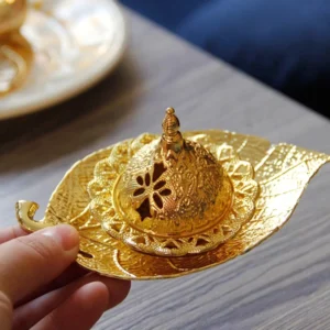 Gold leaf incense burner held by hand, showing intricate carved hollow lid and embossed metal texture on wooden table