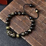 Gold sheen obsidian Pixiu bracelet with Om Mani Padme Hum beads on wooden background, showing glossy golden-black obsidian beads and black adjustable braided cord