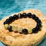 Gold Sheen Obsidian Pixiu Wealth Bracelet with om mani padme hum charm, top-down view on stone with water backdrop showing jet black beads and bright golden accents