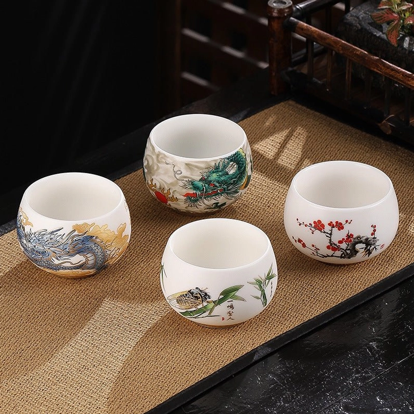 Assorted handcrafted Buddhist tea cups on traditional jute mat showing dragon, bamboo, plum blossom, and crane designs from top view