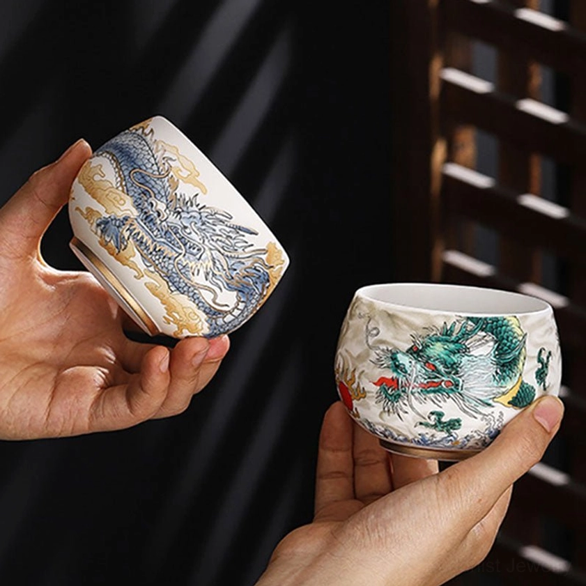 Two handcrafted Buddhist ceramic tea cups held in hands showing blue gilt dragon and green dragon patterns