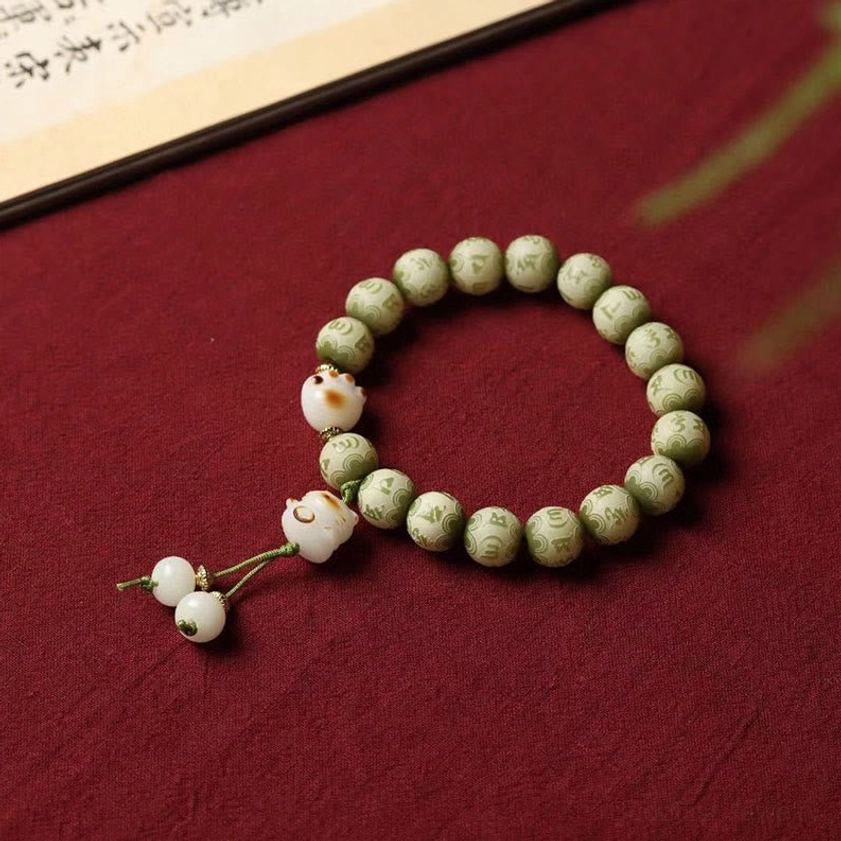 Green Bodhi seed bracelet with engraved Om Mani Padme Hum mantra and white cat paw pendants displayed on dark red fabric