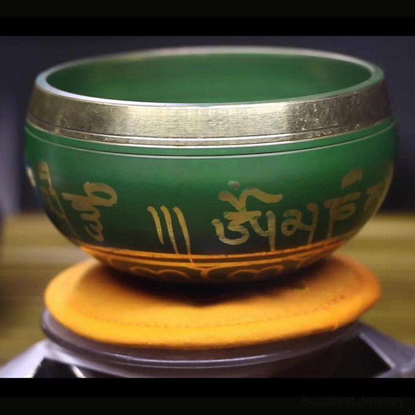 Green Buddhist singing bowl with gold inscriptions and silver metal rim, displayed in side close-up on embroidered orange cushion