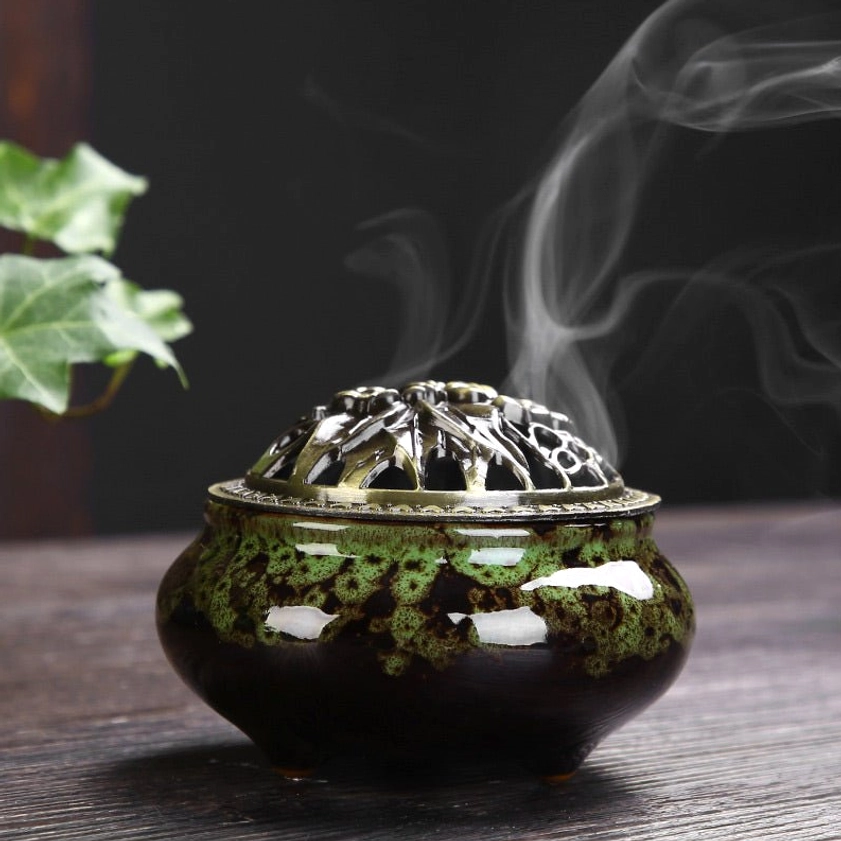 Green glazed ceramic incense holder with bronze floral lid on wooden table with active smoke