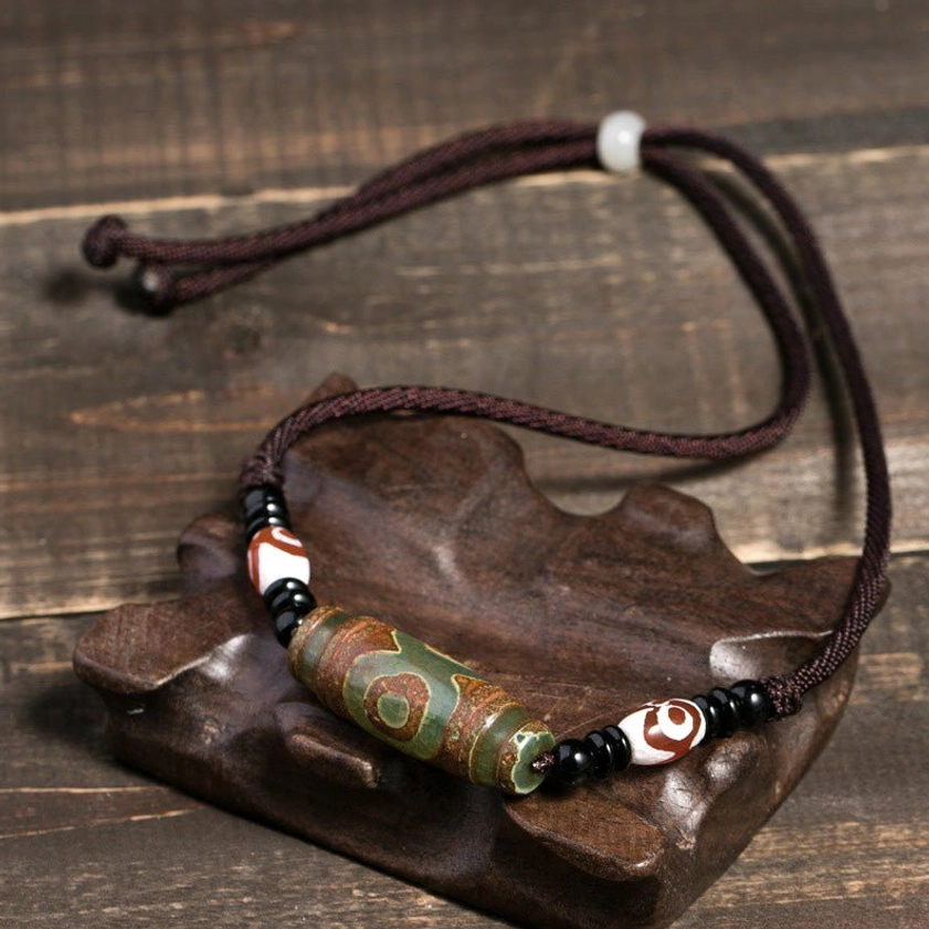 Green Buddhist dzi bead necklace with adjustable brown cord and decorative three-eye accent beads, displayed on wooden stand