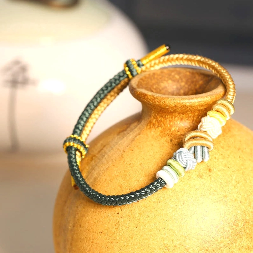 Dark green and gold braided Buddhist lucky bracelet displayed with ceramic vase showing traditional knot details