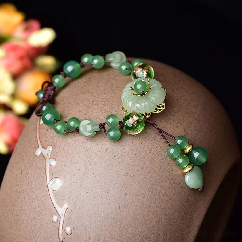 Green jade flower bracelet with gold-trimmed charm and beaded chain displayed on ceramic stand, side overhead view