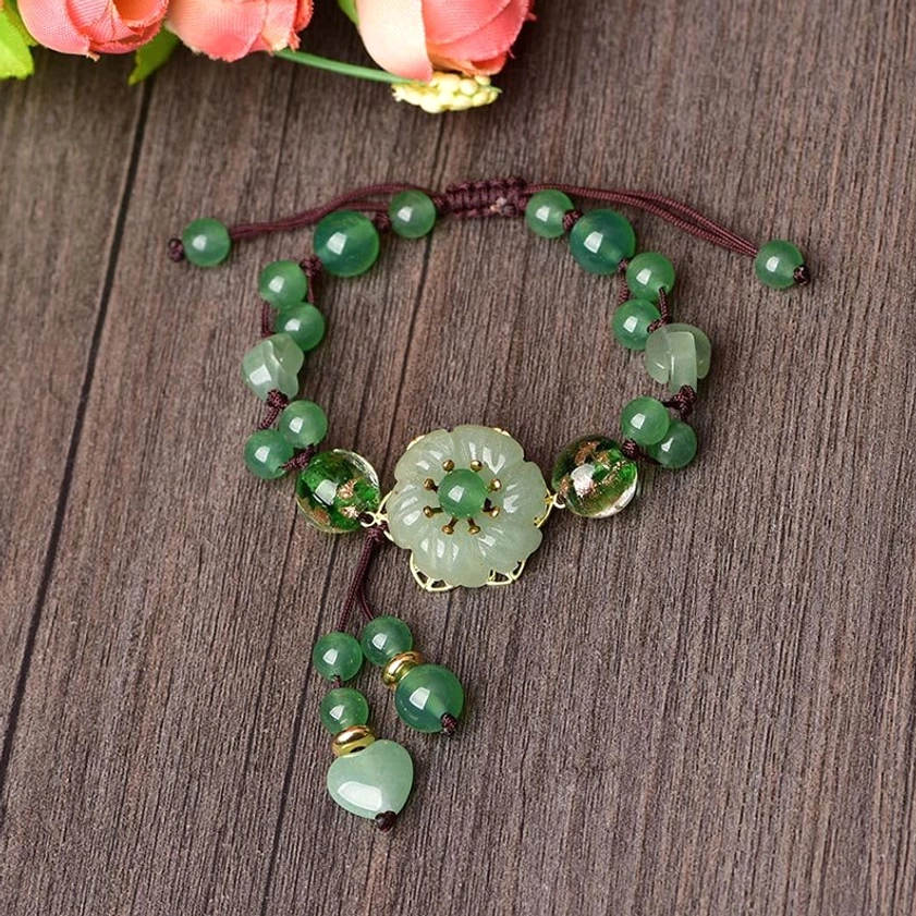 Flat lay of green jade tassel bracelet showing braided brown cord, carved flower charm, and dangling bead pendant on wooden surface