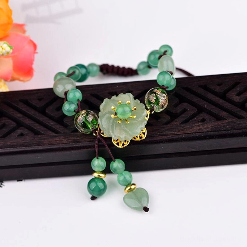Green jade tassel bracelet with gold metal accents and glossy heart pendant on black carved display stand with floral backdrop