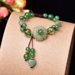 Green jade flower bracelet with dangling charm displayed on light brown stand with floral backdrop, front overhead view