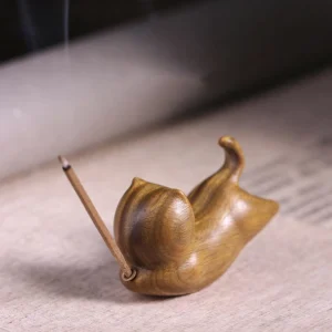Green sandalwood cat-shaped incense holder with burning incense stick, side view showing natural wood grain and rising aromatic smoke on linen desk