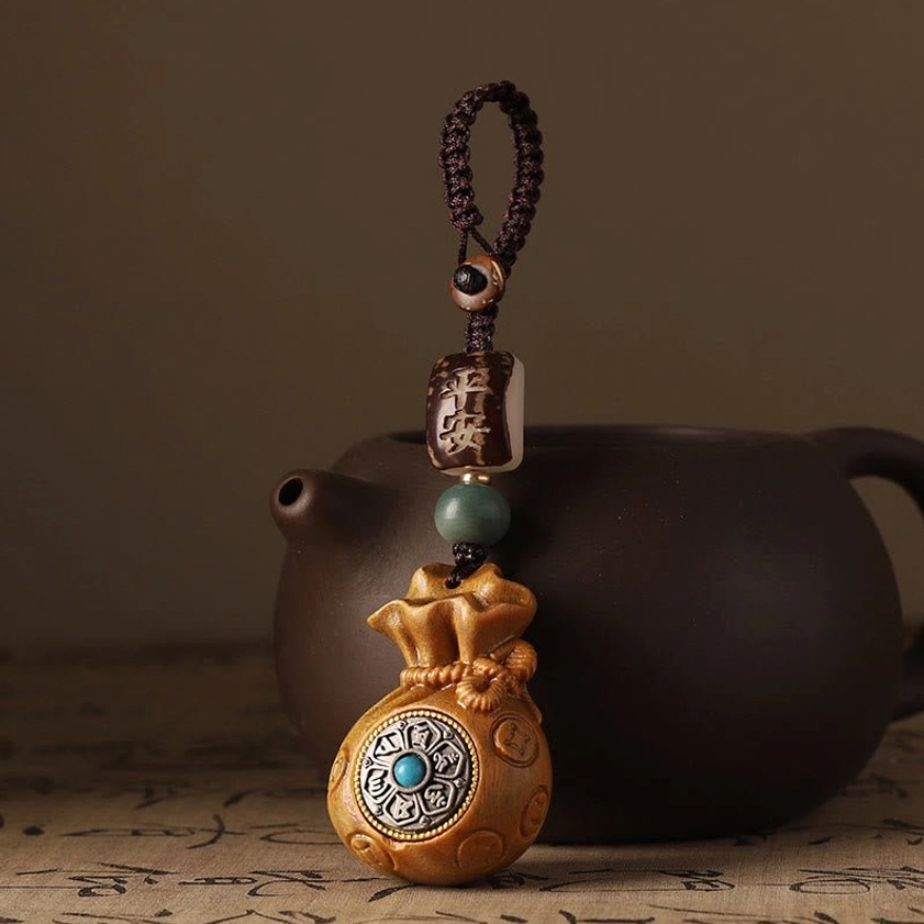 Green sandalwood Buddhist money bag charm with detailed carved money bag shape displayed beside ceramic teapot on calligraphy paper