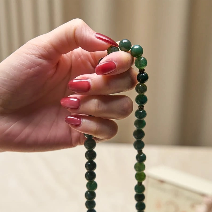 Hand held moss agate Buddhist mala close up view with dangling agate bead strands and gold lotus charm, held by hand with red nails
