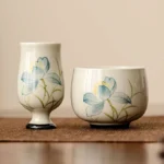 Two hand-painted lotus ceramic tea cups with blue flower design and black base rim on linen placemat - Buddhist mindfulness tea ceremony vessel