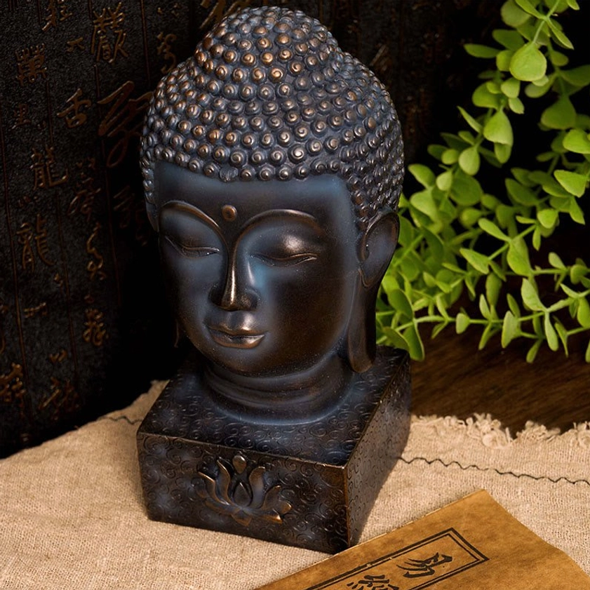 Matte dark bronze Buddha head statue with serene closed-eye expression, lotus engraved base, vintage book display setting