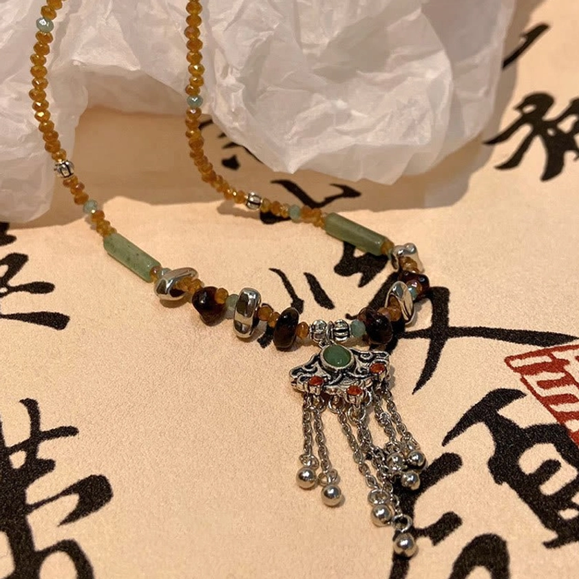 Handcrafted ethnic tassel pendant necklace flat lay displaying mixed earthy beads, silver charm with green enamel inlay, and dangling tassel elements