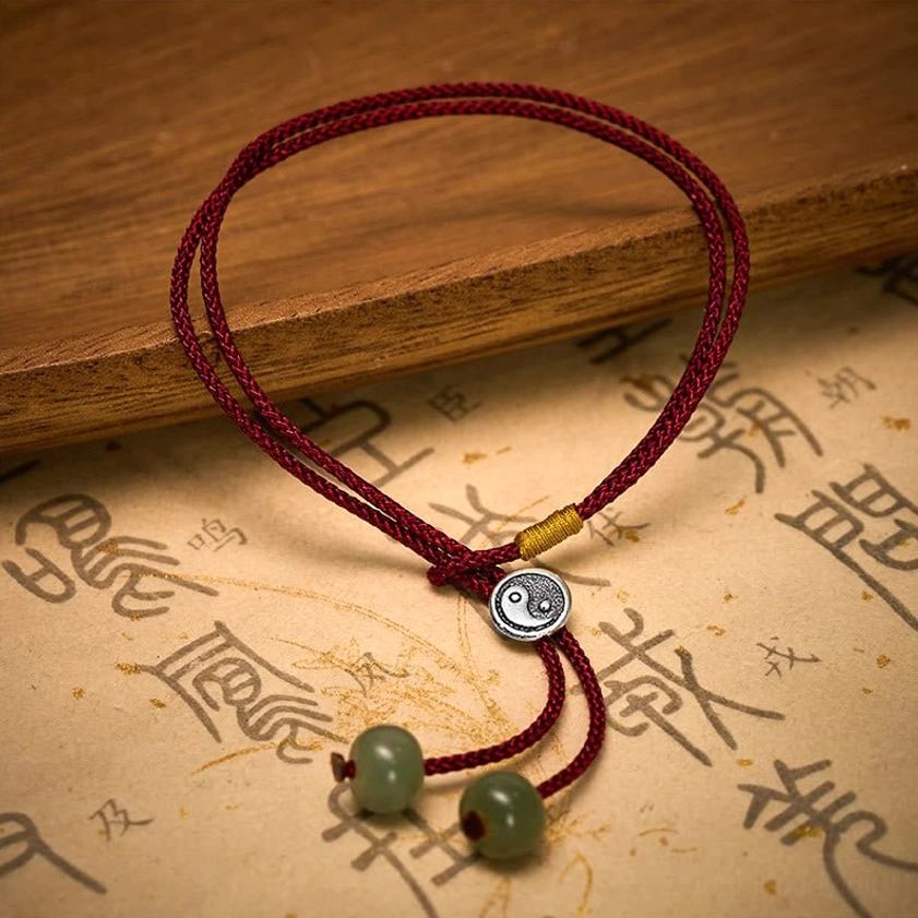 Yin Yang balance symbol bracelet with braided red cord, sterling silver pendant, green beads, and gold cord accent fastener, styled on parchment and wood