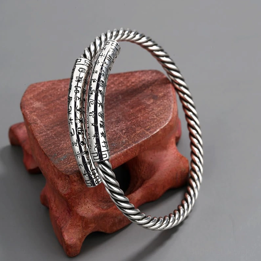Double twisted copper Heart Sutra cuff bangles with open design and engraved Buddhist text, front three-quarter view on red wooden display stand