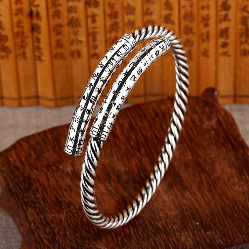 Hand-engraved Heart Sutra copper cuff bangle in antique silver tone with twisted rope texture, shown from three-quarter oblique angle on wooden display base