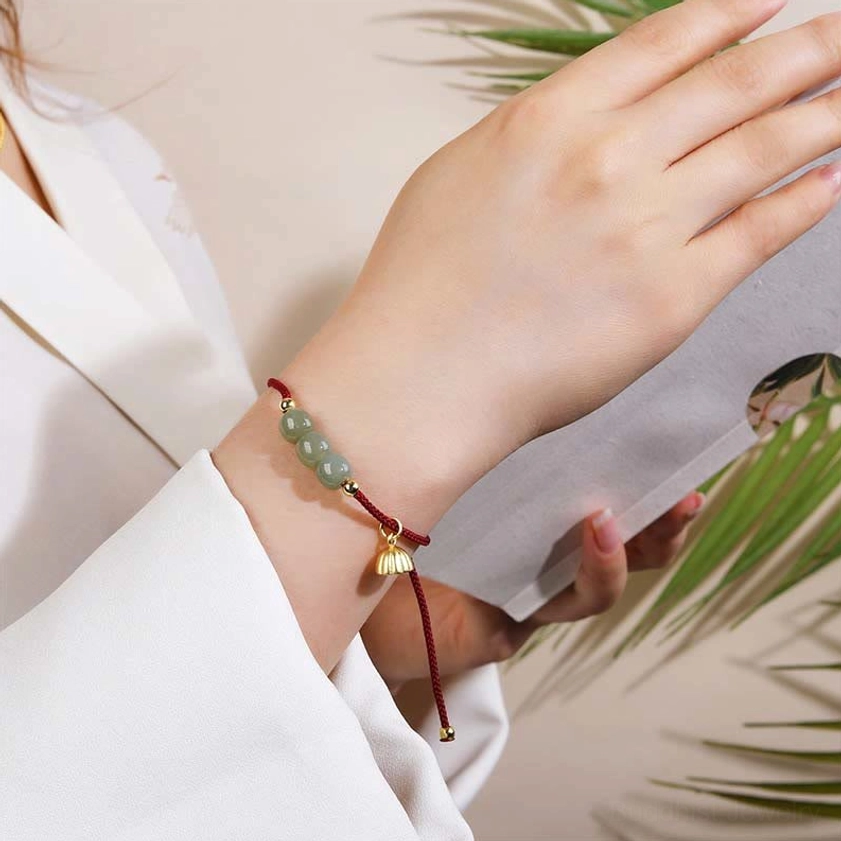 Lifestyle photograph of jade prosperity bracelet worn on wrist with red woven cord and gold lotus charm, styled in indoor reading environment with green plants
