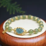 Hetian jade lotus bracelet with gold-plated silver charms displayed on wooden base with green foliage, showing light green jade beads and dangling prosperity tag