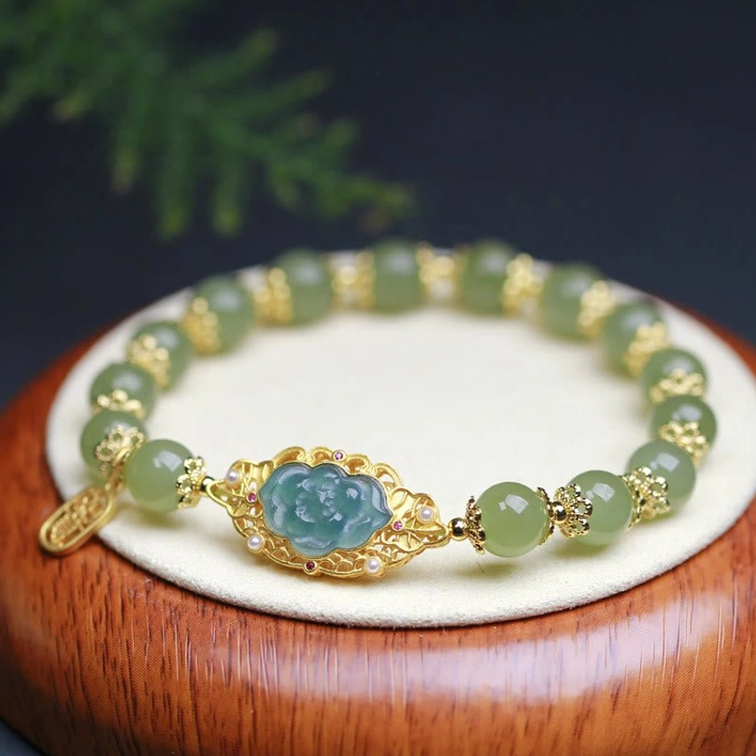 Hetian jade lotus bracelet with gold-plated silver charms displayed on wooden base with green foliage, showing light green jade beads and dangling prosperity tag