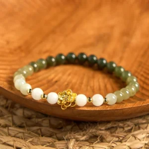 Hetian jade lotus bracelet with white to green gradient beads and gold lotus charm, displayed on wooden tray with woven mat background