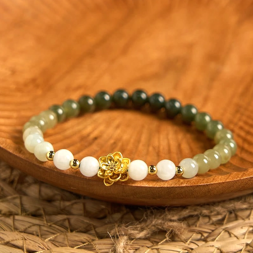 Hetian jade lotus bracelet with white to green gradient beads and gold lotus charm, displayed on wooden tray with woven mat background
