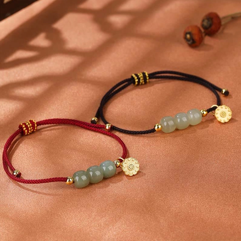 Hetian jade lotus prosperity bracelet with red woven cord and gold charm displayed on warm fabric with dried botanical elements, showcasing the handcrafted adjustable design