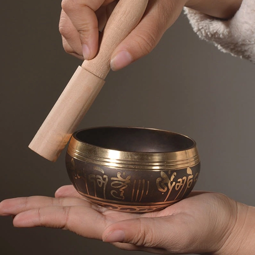 Hands demonstrating proper technique for striking and playing Buddhist singing bowl with smooth wooden mallet