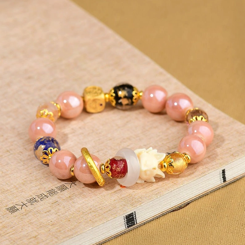 Buddhist prayer bead bracelet with nine-tailed fox charm, pink liuli glass beads, gold accents, and decorative blue and red beads, arranged on ancient Chinese calligraphy book