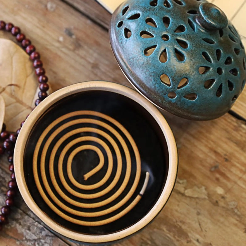 Top-down view of ceramic incense burner with separated teal lid and dark brown base holding spiral coil incense, displayed with prayer beads and dried foliage