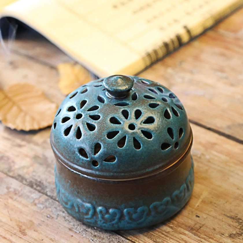 Front side view of closed ceramic incense burner with round finial, floral hollow carved lid, and faint smoke rising from gaps on rustic wooden table