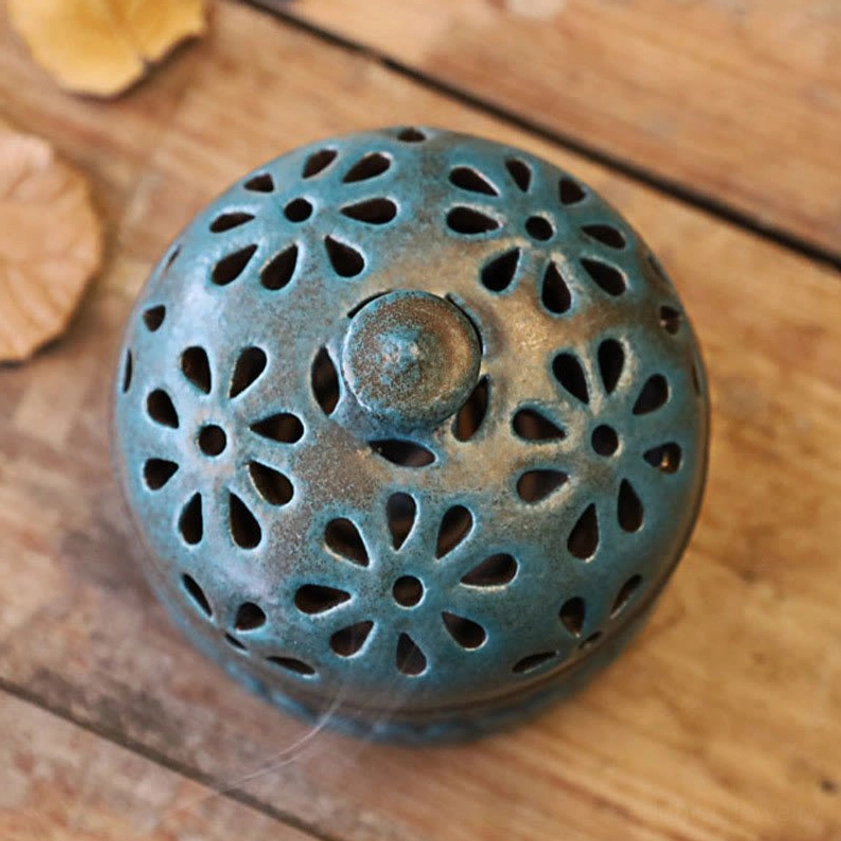 Close-up overhead shot of incense burner lid showcasing teal-blue floral hollow carvings with round ceramic knob on dried leaf backdrop