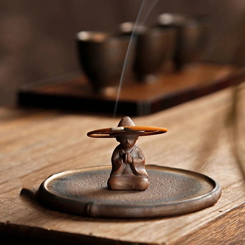 Overhead view of burning incense stick in ceramic monk incense holder on tea table with tea cups and curling fragrant smoke
