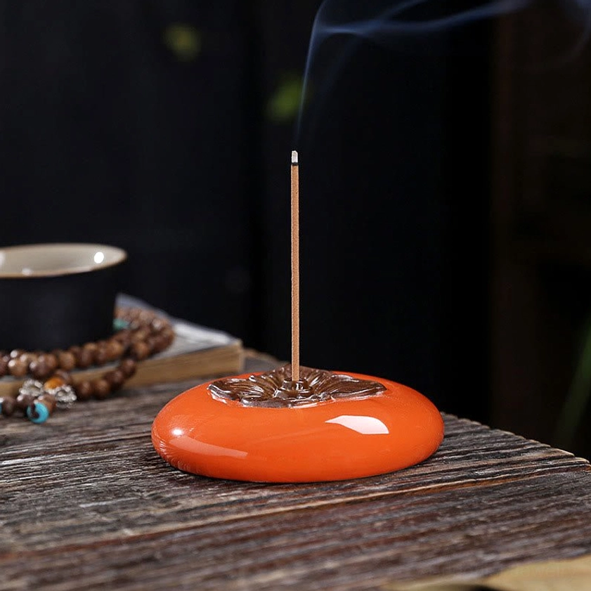 Burning persimmon ceramic incense holder on wooden table with prayer beads and tea bowl nearby in peaceful tea leisure scene