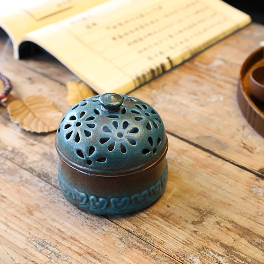 Closed ceramic incense burner showing two-tone finish with embossed base trim and hollow floral carved lid, styled on wooden table with book and meditation accessories