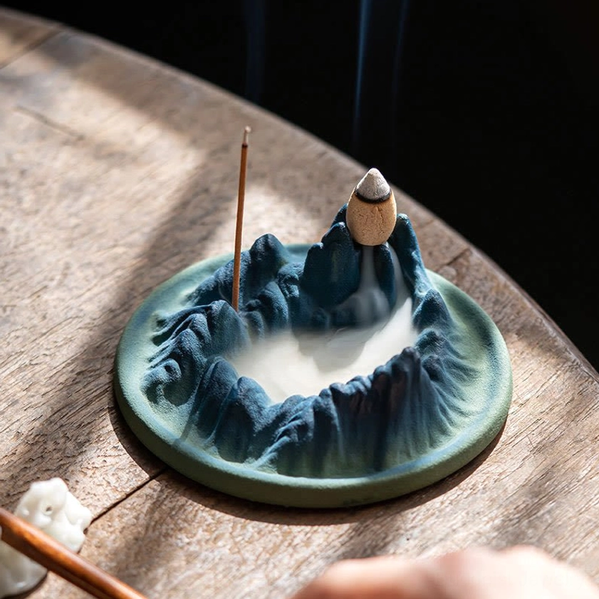 Buddhist ceramic backflow incense burner on rustic wooden round table with incense stick burning and aromatic smoke flowing downward