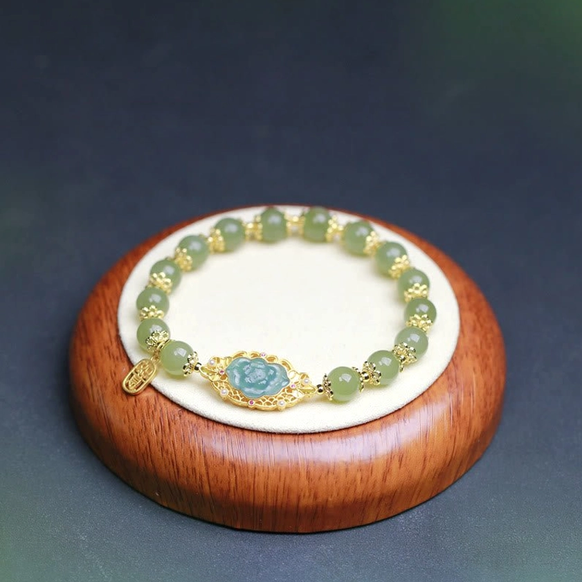 Hetian jade lotus luck bracelet on wooden round base with dark background, showing polished jade beads, gold filigree lotus charm, and dangling prosperity tag