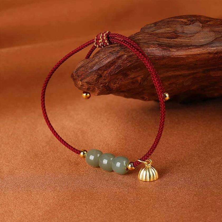 Close-up minimalist view of hetian jade bracelet with red adjustable cord, tiny gold beads, and lotus pod charm resting on warm brown wooden surface
