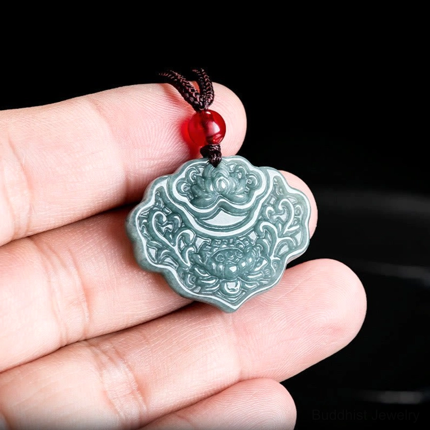 Close-up handheld jade lotus pendant held between fingers, showing natural jade texture, brown rope cord, and red protective bead up close