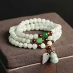Natural jade pixiu buddha bracelet with multi-strand design resting on wooden jewelry box, showing light green beads with jade pendants and red spacer accents