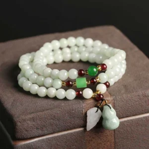 Natural jade pixiu buddha bracelet with multi-strand design resting on wooden jewelry box, showing light green beads with jade pendants and red spacer accents