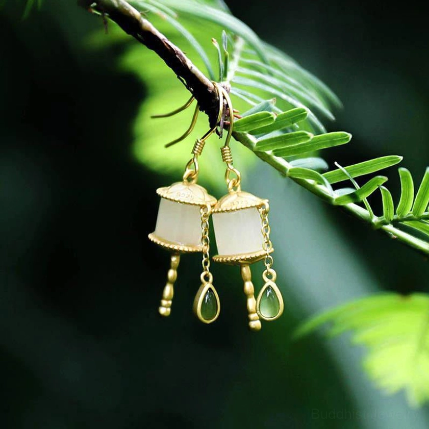 Buddhist jade prayer wheel drop earrings front view with tassel-style dangle design, dark green foliage background, gold silver jade