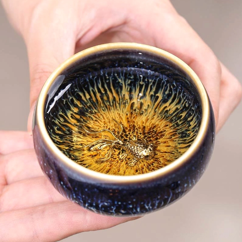 Held Jian Zhan ceramic tea cup with koi fish ornament, eye-level view showing dark exterior and gold-rimmed interior glaze