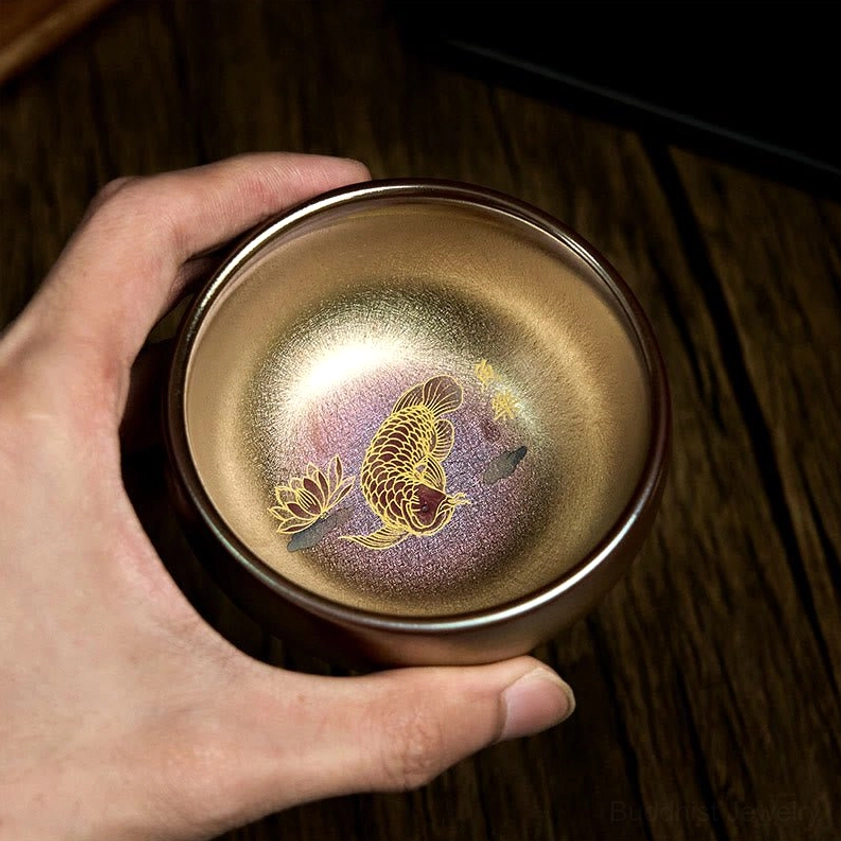 Hand-held side-top view of handmade Jianzhan tea cup showing metallic bronze-gold glaze and inner koi lotus design